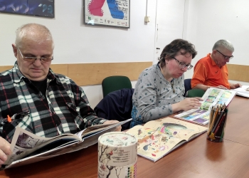 Uczestnicy oglądają i czytają komiksy rozłożone na stole. Trzy osoby są skupione na przeglądaniu komiksów. 