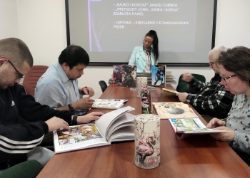 Na zdjęciu widać grupę osób siedzących przy długim stole.Uczestnicy przeglądają i czytają komiksy. Na końcu stołu stoi kobieta prowadząca wydarzenie. Za nią wyświetlana jest prezentacja dotycząca komiksów — na slajdzie można odczytać m.in. tytuły „Kajko i Kokosz”, „Przygody Jonki, Jonka i Kleksa” oraz wzmiankę o Japonii i wielopiętrowych księgarniach z komiksami. Na stole ustawiono kilka komiksów na ekspozycji, w tym album z Supermanem. 