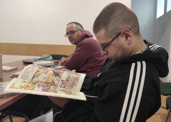 Na zdjęciu widać dwóch mężczyzn siedzących przy stole podczas spotkania poświęconego komiksom. Jeden z nich z dużym skupieniem czyta otwarty komiks, a drugi obserwuje go z drugiego końca stołu.