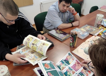 Na zdjęciu kilka osób siedzi przy stole i przegląda różne komiksy oraz książki obrazkowe. Uczestnicy wyglądają na skupionych i zainteresowanych lekturą, a na stole widać kolorowe albumy, kredki oraz materiały związane z tematyką komiksową.