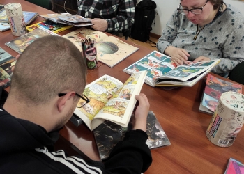Na zdjęciu uczestnicy spotkania siedzą przy stole i z zainteresowaniem czytają kolorowe komiksy oraz albumy ilustrowane.
