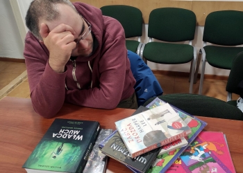 Na zdjęciu mężczyzna siedzi przy stole. Na stole leżą różnorodne komiksy.